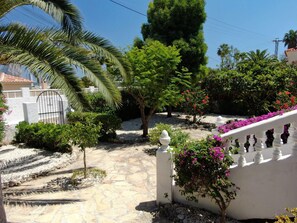 Ein gepflasterter Weg führt zu einem Gebäude mit weißen Wänden und einem verschlossenen Eingang, umgeben von üppiger Vegetation und blühenden Pflanzen.