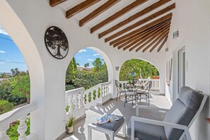 Uma área de estar coberta ao ar livre, com mesa, cadeiras e um sofá, e uma silhueta de árvore decorativa na parede.