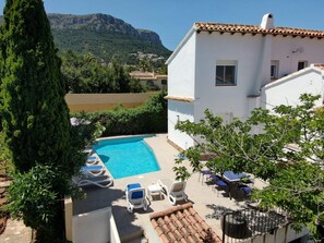 A pool area with lounge chairs, a white building with a tiled roof, and a mountainous landscape in the background. - Villa San Marcos - PlusHolidays (Calpe)