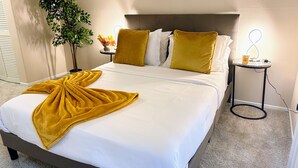 Un lit bien fait, agrémenté d’un plaid jaune, de draps blancs et de deux coussins assortis. Une petite table avec une lampe et une plante se trouve à côté du lit.