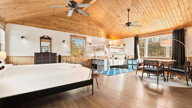 A spacious room with a large bed, wooden ceiling, two ceiling fans, a desk with chairs, and a window with a view of trees.