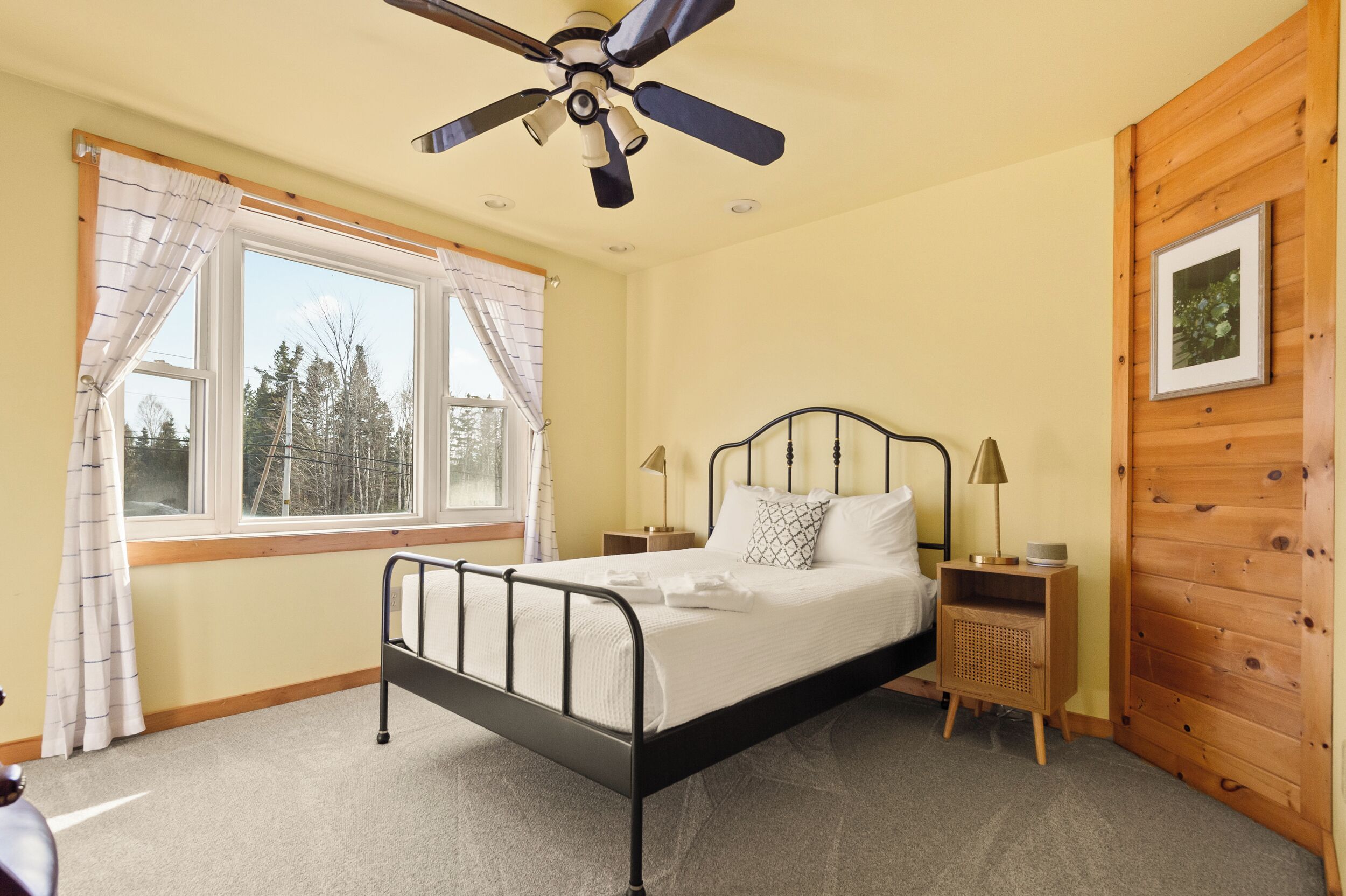Un dormitorio con cama, ventilador de techo, ventana con cortinas, mesita de noche con lámpara y panel de pared de madera.
