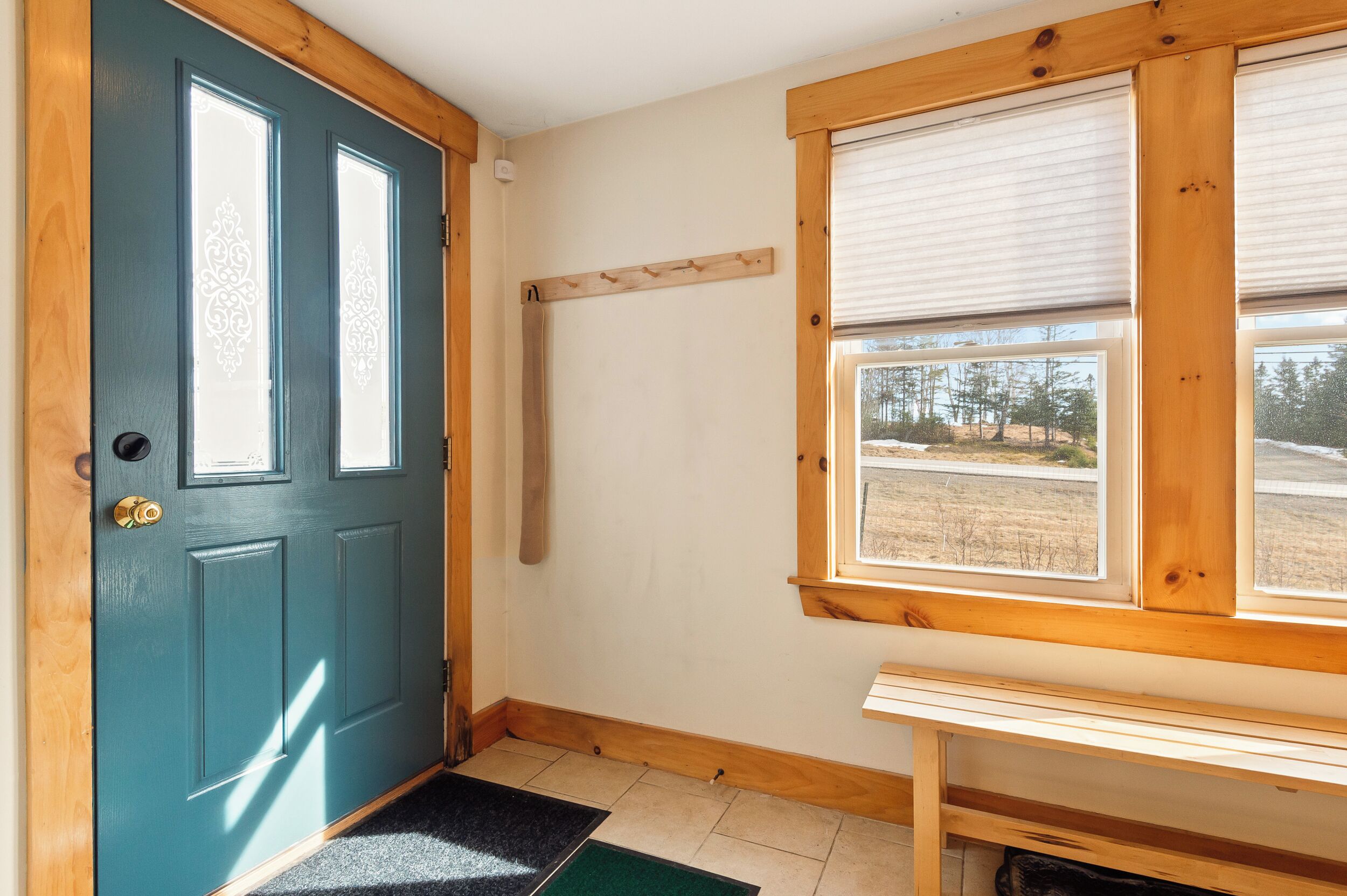 Una stanza con una porta blu, una panca di legno, una finestra vista alberi e un muro bianco.