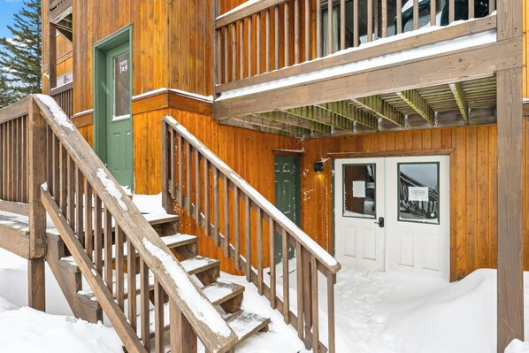 A wooden building with a green door, a staircase with a wooden railing, and a snow-covered ground.