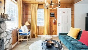 A cozy cabin interior with a stone fireplace, a wooden ceiling, a green sofa, and a dining table.