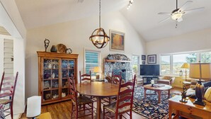 A spacious living room with a wooden dining table, red chairs, a stone fireplace, and a large window offering a view of the outdoors.