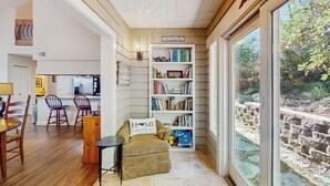 A cozy living room with a bookshelf, a dining area with wooden chairs, and a view of a garden through a large window.