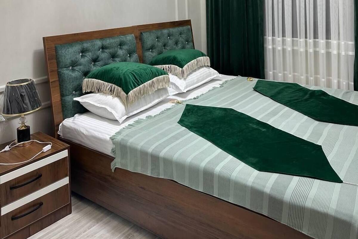 A bedroom with a wooden bed frame, green and white bedding, a bedside table with a lamp, and a window with sheer curtains.