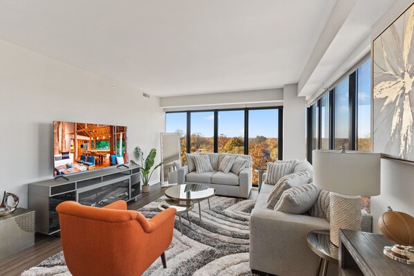 A modern living room with a large TV, grey sofas, an orange armchair, and a view of autumn trees through large windows. - "Hope of Evanston" 180°view, 2BDR +2Bath Urban luxury (Evanston)