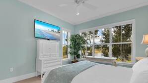 Ein Schlafzimmer mit Bett, Wandfernseher, Fenster mit Blick auf Bäume und Deckenventilator.