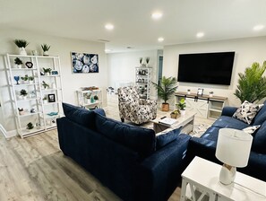 A modern living room with a dark blue sofa, a patterned armchair, and a white coffee table. There are shelves with plants and decorative items, a large flat-screen TV, and a wooden floor.