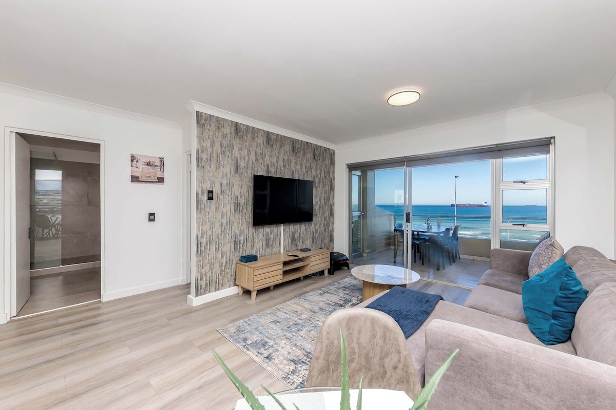 Una sala de estar moderna con un televisor de pantalla plana, un sofá y un comedor con vistas al mar.