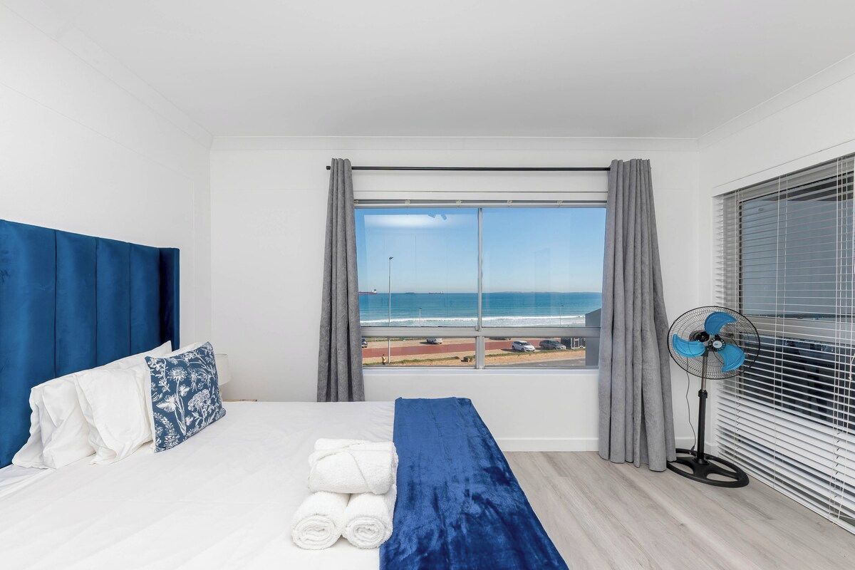 A hotel room with a bed, a view of the beach, a standing fan, and a window with curtains.