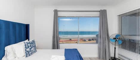 Une chambre d’hôtel avec un lit, une vue sur la plage, un ventilateur sur pied et une fenêtre avec des rideaux.