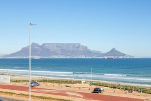 A coastal road with a mountain in the background, a beach, and a clear sky.