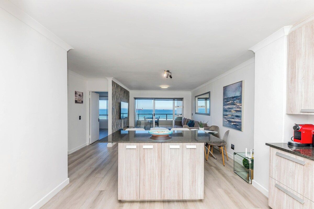 A modern kitchen with a central island, large windows, and a view of the sea.
