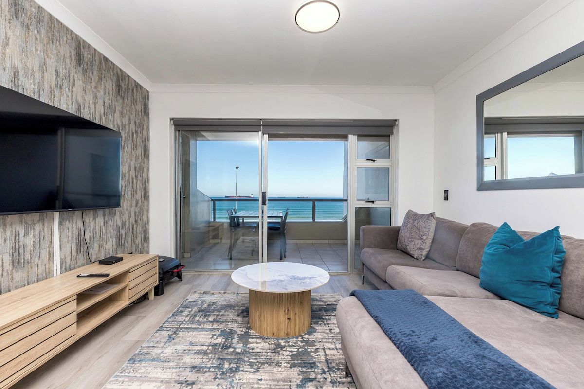 Un salon moderne avec un téléviseur à écran plat, un meuble de rangement en bois, une table basse ronde, un canapé beige agrémenté de coussins turquoise et une vue sur la mer grâce à des portes vitrées.