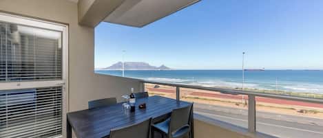 A balcony with a table and chairs, overlooking a coastal road and ocean.
