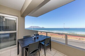 Een balkon met een tafel en stoelen, uitkijkend op een kustweg en de oceaan.