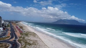 A coastal road with a beach and ocean on the right, buildings on the left, and a mountain in the distance. - Seaside Living on Kite Beach, Blouberg (Cape Town)