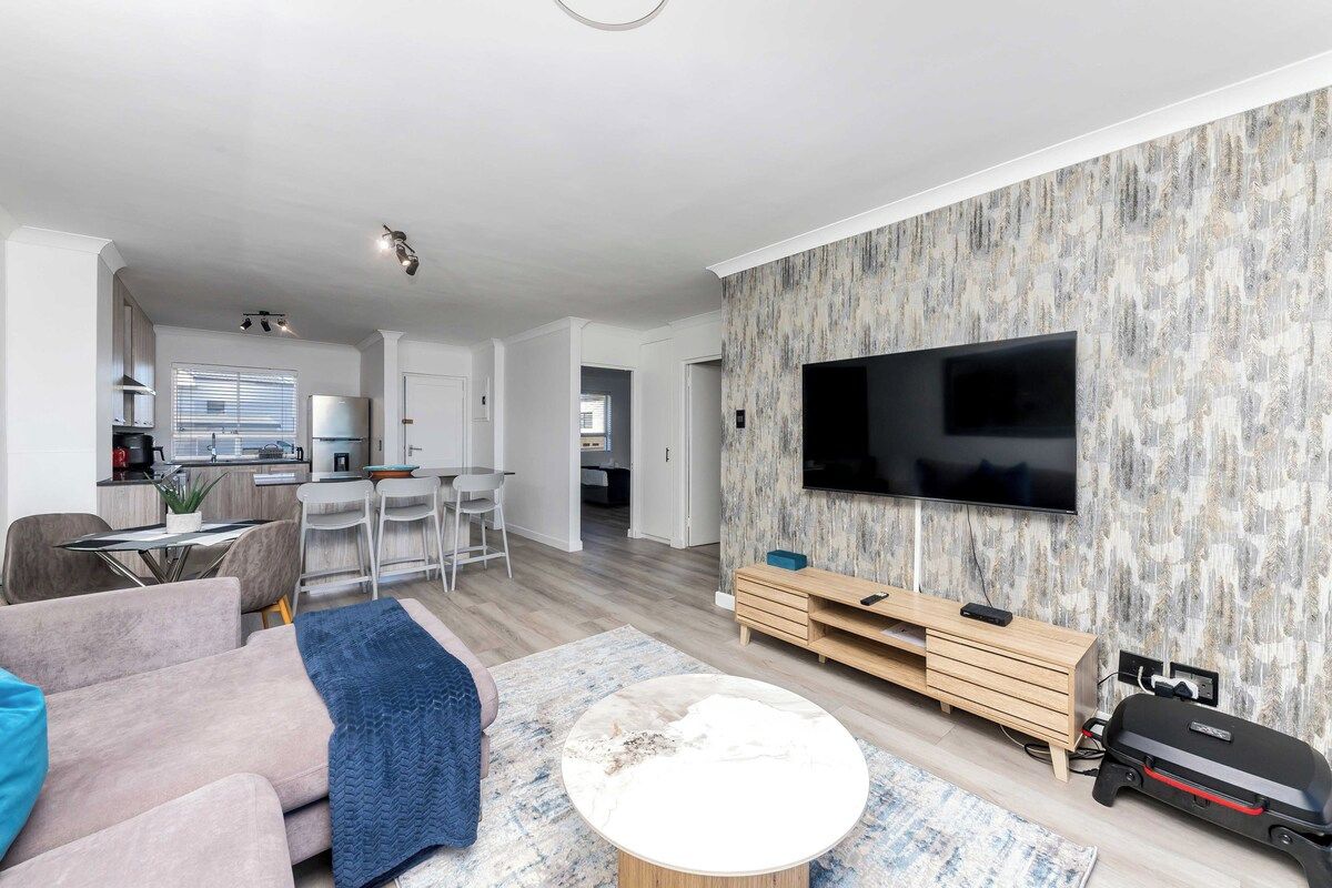 A modern living room with a flat-screen TV mounted on the wall, a sofa, a coffee table, and a dining area with a kitchen in the background.
