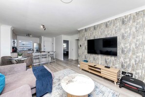 A modern living room with a flat-screen TV mounted on the wall, a sofa, a coffee table, and a dining area with a kitchen in the background. - Seaside Living on Kite Beach, Blouberg (Cape Town)