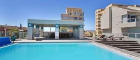 A modern hotel with a swimming pool, a covered area, and a multi-story building in the background.