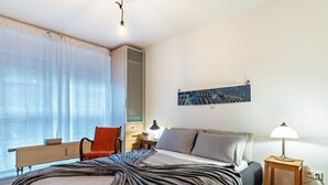 Um quarto com uma cama, uma cadeira, uma mesa de cabeceira, uma luminária, um quadro e uma janela com cortinas.