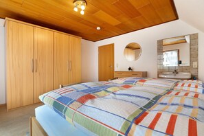 Une chambre à coucher avec une armoire en bois, un lit recouvert d’une couette aux couleurs vives, un miroir rond et une salle de bain avec un lavabo.