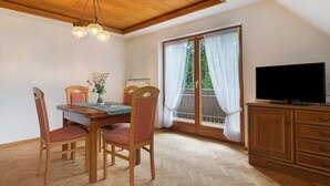 Uma sala de jantar com cadeiras de madeira, uma mesa, um lustre, uma televisão sobre um armário de madeira e uma janela com cortinas.