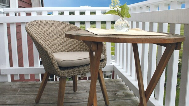 A wicker chair with a cushion on a wooden deck, a small round table with a vase and a plant, and a white picket fence. - Apartment 'Ferienwohnung Traumblick' with Balcony and Wi-Fi (Reesdorf)