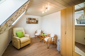 A room with a sloped ceiling, a sofa, two wicker chairs, a small table, a wooden wardrobe, and a window.