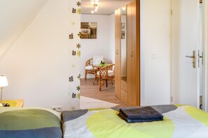 A bedroom with a bed, a wooden wardrobe, a dining area with a table and chairs, and a wall with a floral pattern.