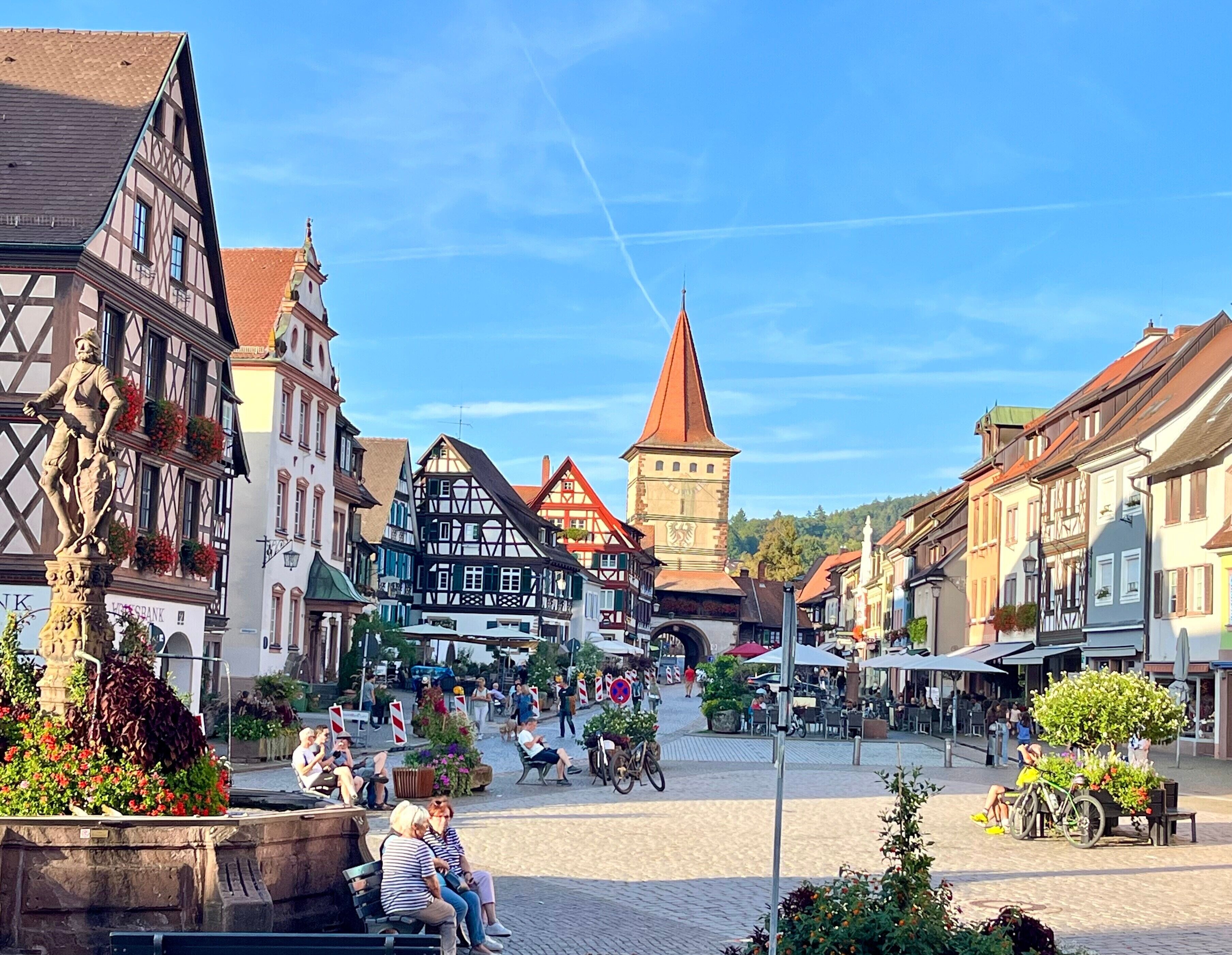 Uma praça histórica com edifícios tradicionais em estilo de enxaimel, uma torre imponente e uma estátua.