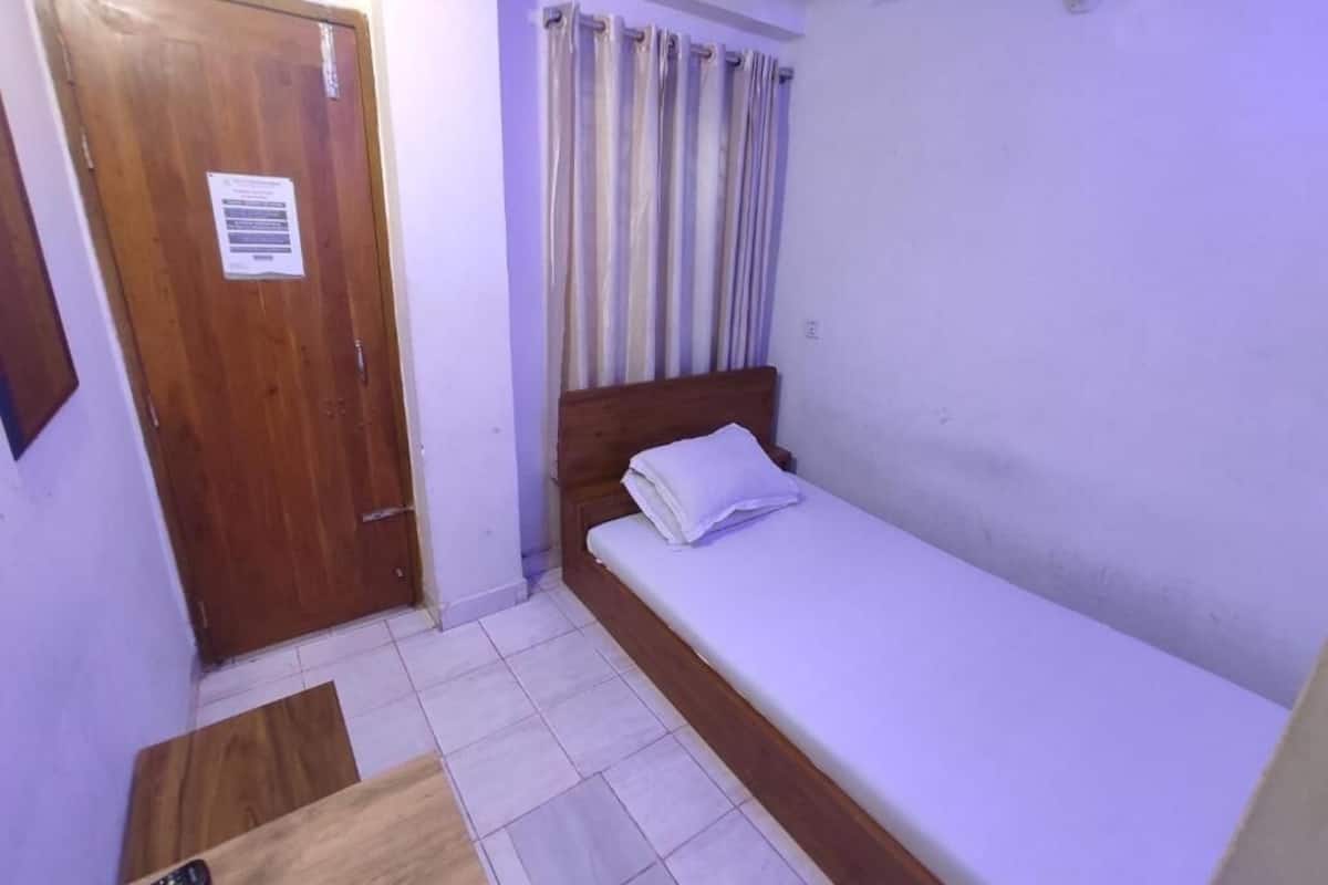 Une petite chambre avec un lit simple, une porte en bois, un rideau et une télécommande posée sur une table en bois.