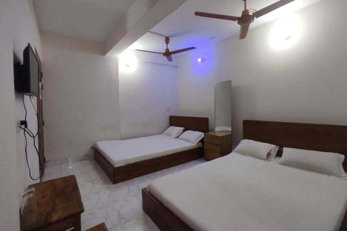 Une chambre avec deux lits simples, une table de chevet en bois, un téléviseur fixé au mur et deux ventilateurs de plafond.