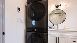 A modern bathroom with a black washing machine, a round mirror, and a white vanity with cabinets. - Cozy, Bran New Private fully furnished studio downtown Chilliwack.  (Chilliwack)