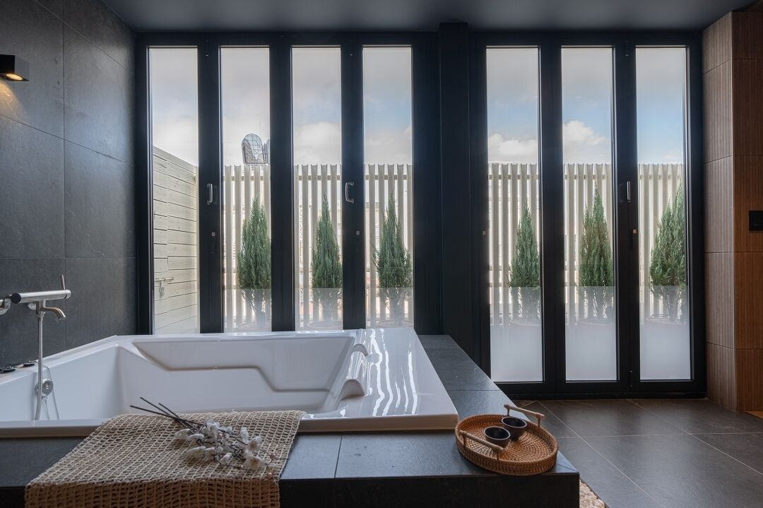 A modern bathroom with a large bathtub, a window with a view of a wooden fence and trees, and a small wicker basket on the floor.