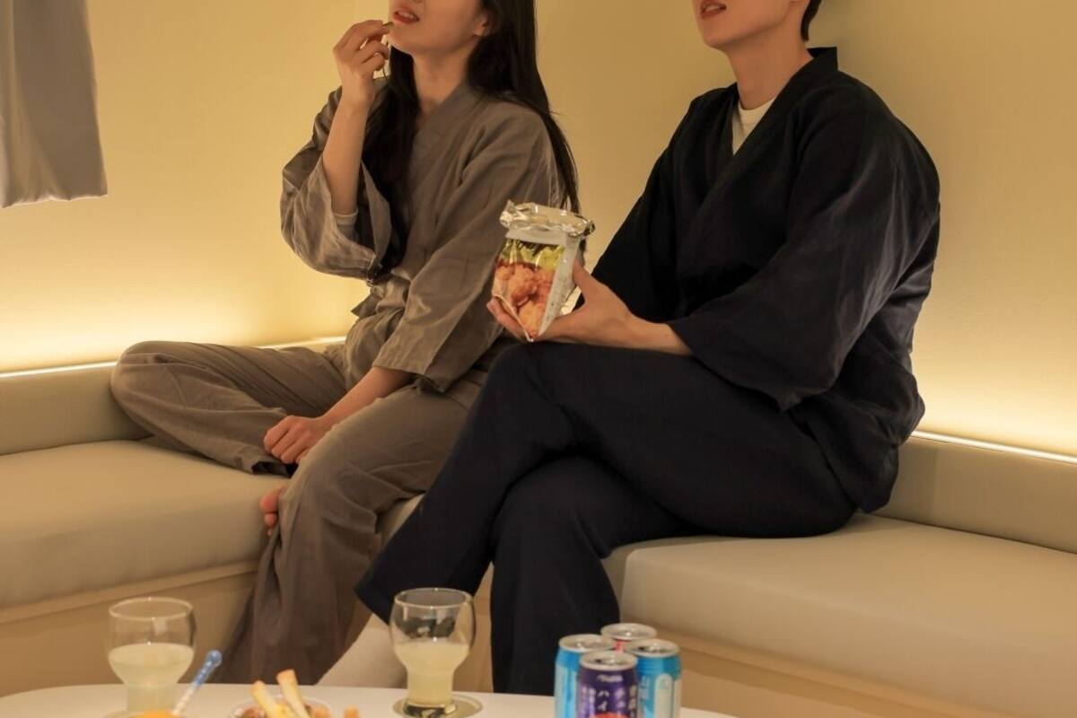 Two people sitting on a sofa with a small table in front of them. The table has a bowl of food, a glass of drink, and a can of soda.