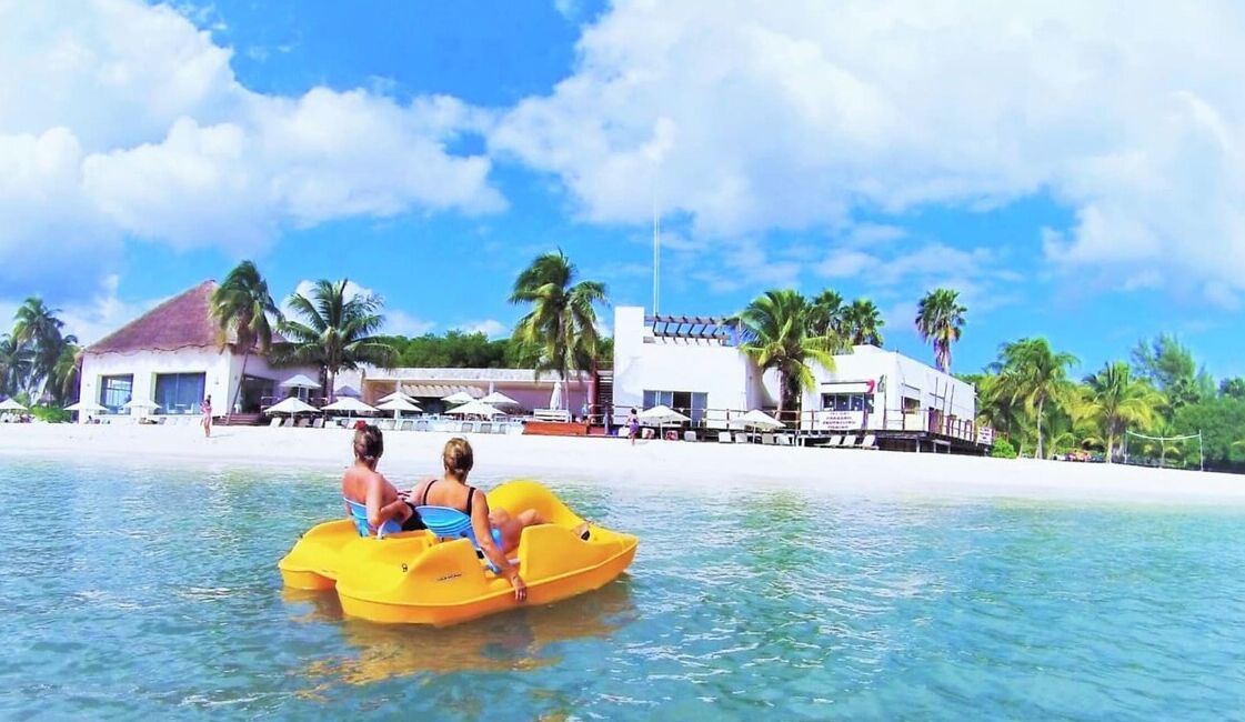 Une scène de plage avec un radeau jaune sur l’eau, des palmiers et un bâtiment avec un toit rouge en arrière-plan.