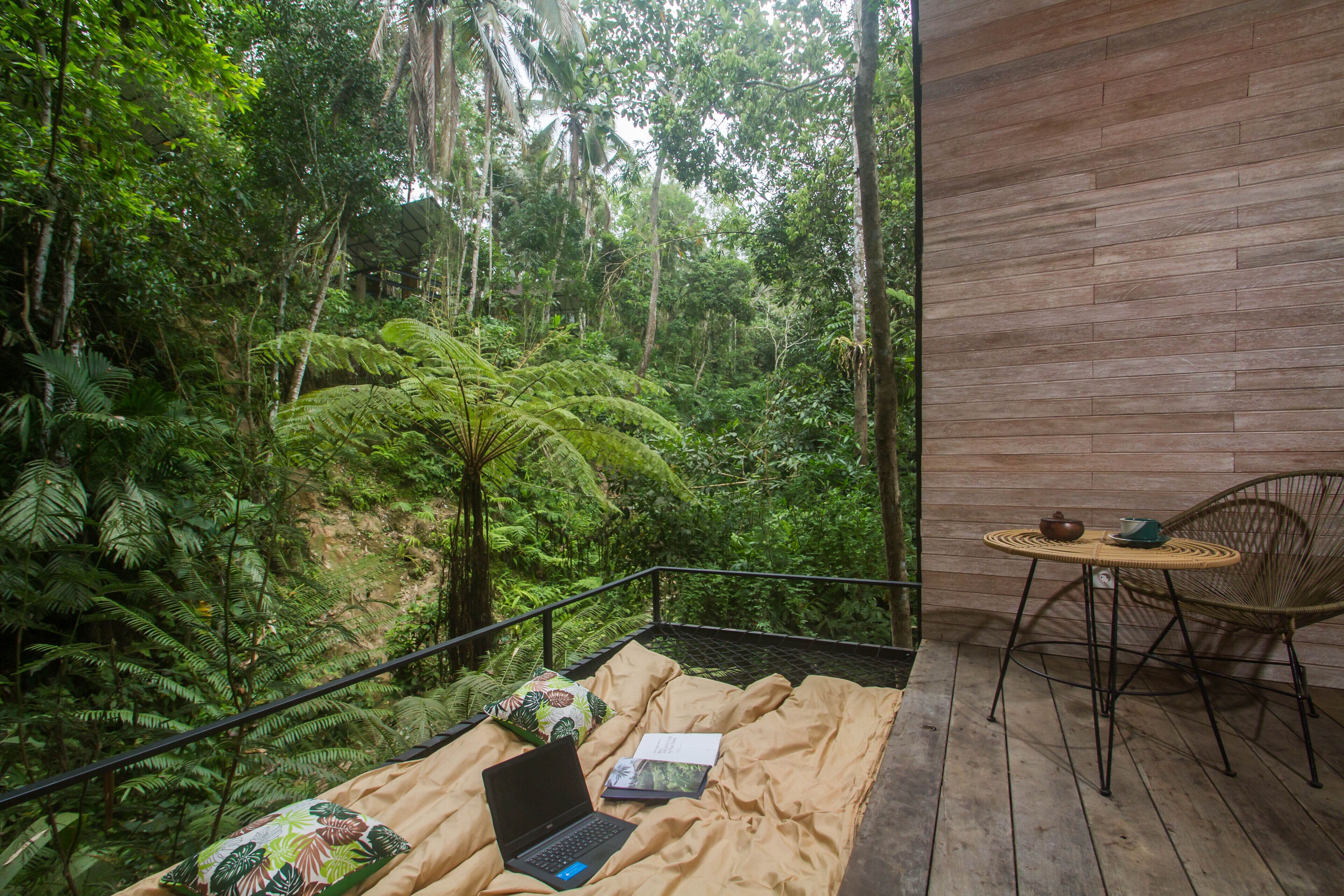 Una habitación con una cama, una computadora portátil y una mesa con una silla. El suelo es de madera y se ve un bosque frondoso.