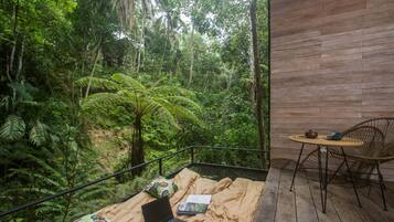 Una habitación con una cama, una computadora portátil y una mesa con una silla. El suelo es de madera y se ve un bosque frondoso.