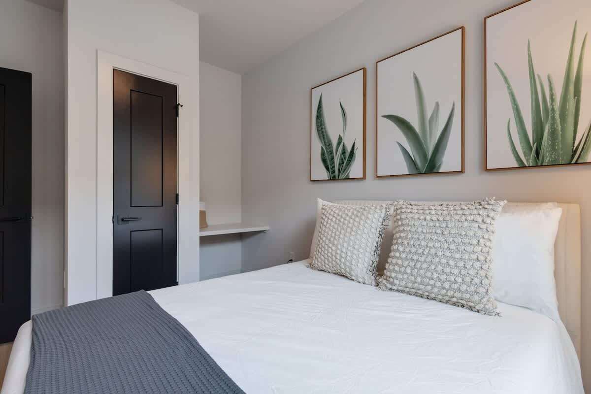 A bedroom with a bed, pillows, a door, and wall-mounted plant art.