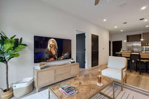 A modern living room with a flat-screen TV, a coffee table with books, a white sofa, a dining area with a kitchen in the background.