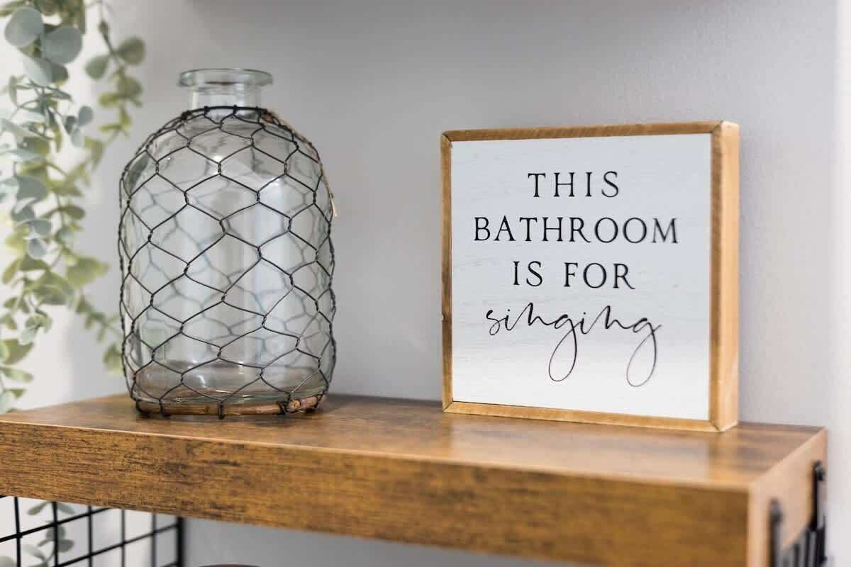 A bathroom sign with the text 'THIS BATHROOM IS FOR singing' and a glass bottle with a wire mesh design on a wooden shelf.