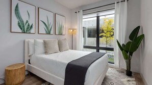 A modern bedroom with a bed, a bedside lamp, a large window with curtains, and a potted plant. - Savanna-Odyssey at the Park- Pool & Full Gym (Nashville)
