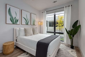 A modern bedroom with a bed, a bedside lamp, a large window with curtains, and a potted plant.