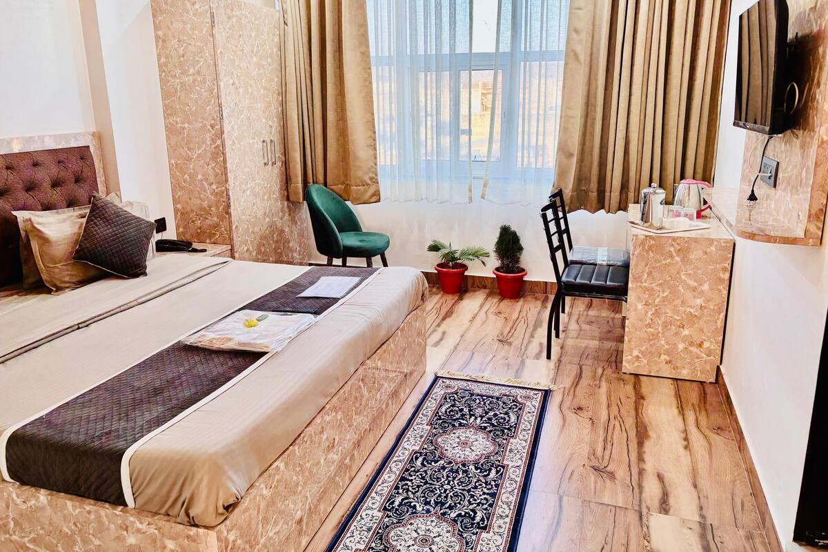 Ein Hotelzimmer mit einem großen Bett, einem Schreibtisch, einem Sessel, zwei Blumentöpfen und einem an der Wand befestigten Fernseher.