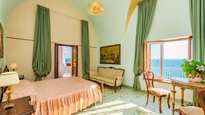 Una habitación con cama, sofá, mesa con sillas, candelabro, un cuadro y vista al mar.