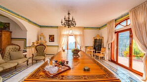 Una habitación amplia con una gran mesa de madera, sillas ornamentadas, una araña y vistas a través de un ventanal.